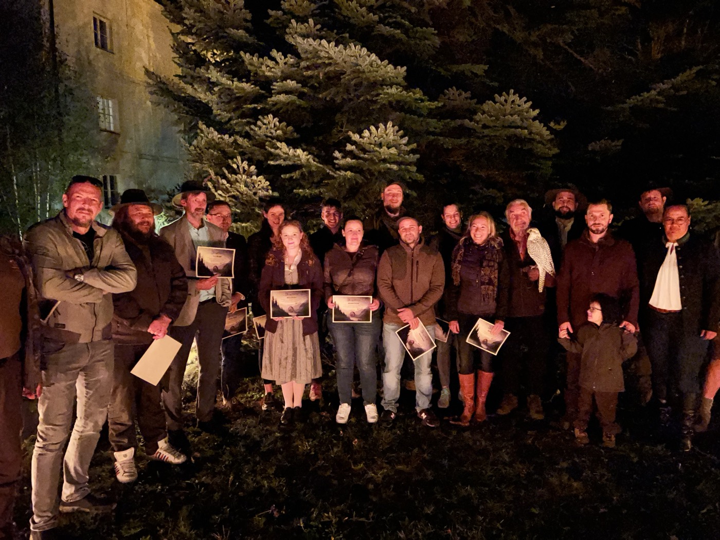 Grosse Gruppe Absolventen posiert abends vor Tanne mit gerahmten Bildern bei Abschlussfeier der Jagdschule